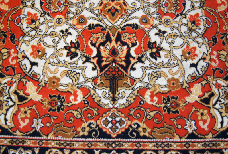 Close-up view of an ornate patterned rug. The rug is predominantly red and white with black and orange accents.