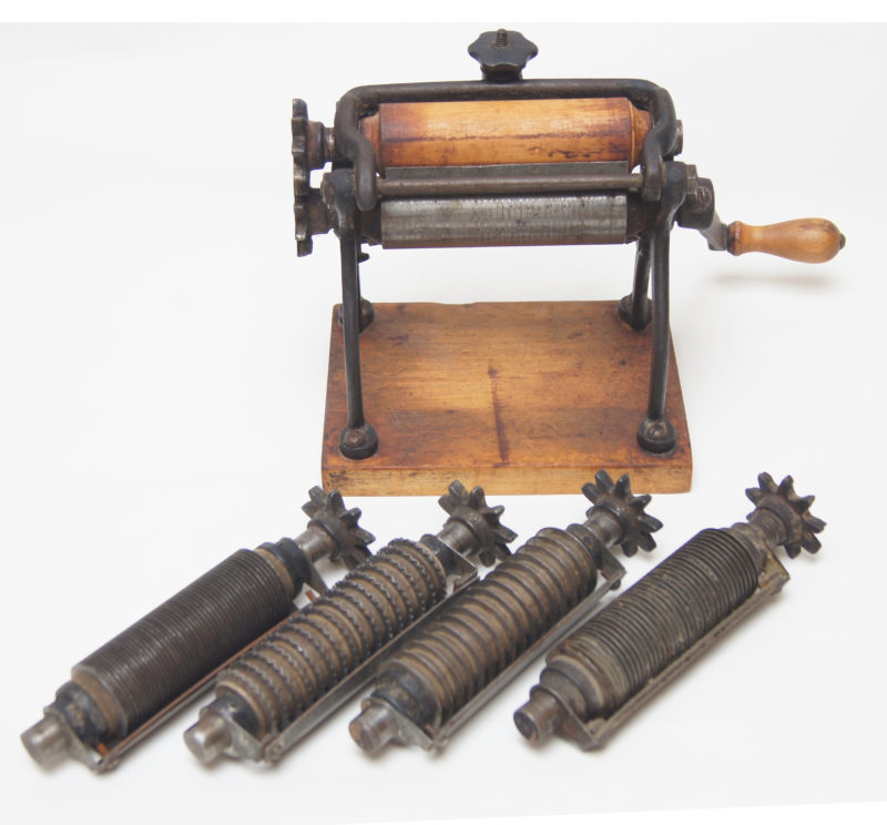 Front view of an 1896 pasta machine. A metal machine sits on top of a wooden square. The machine has a crank with a wooden handle.