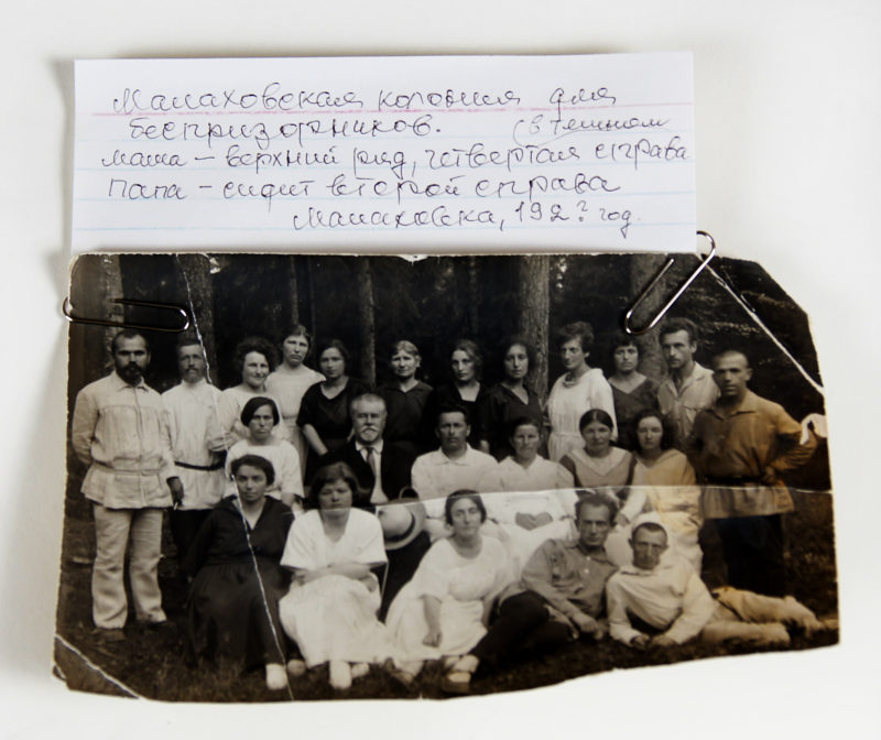 Front view of a black and white photograph paperclipped to an index card with a note hand-written in Russian. The photograph features three rows of men and women posing for the camera in front of trees.