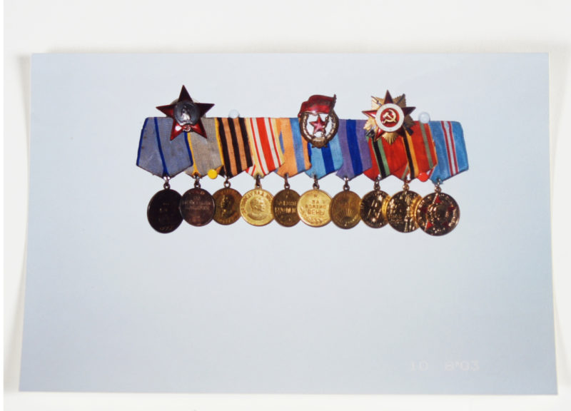 Front view of a photo of ten medals with colorful ribbons lined-up and hanging on a white wall.