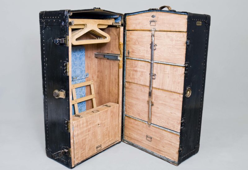 A black leather trunk stands open on its side to reveal a travel wardrobe. The interior is wooden and features various drawers and built-in hangers.