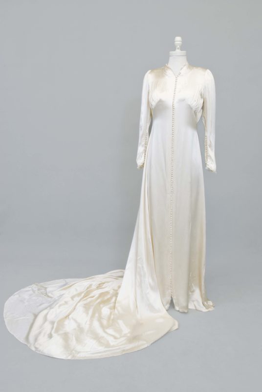 Frontal view of a white satin wedding gown and train. The gown is fitted to a dress form and is standing upright.