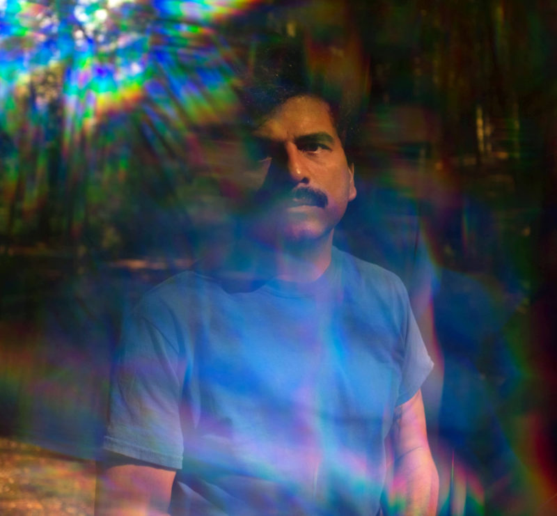 Man with a black mustache looks at the camera. He is slightly obscured by refracted light in the upper left corner.