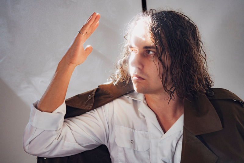 A man with long, brown hair wears a white shirt and brown jacket. He is blocking the sun from his face with his right hand.