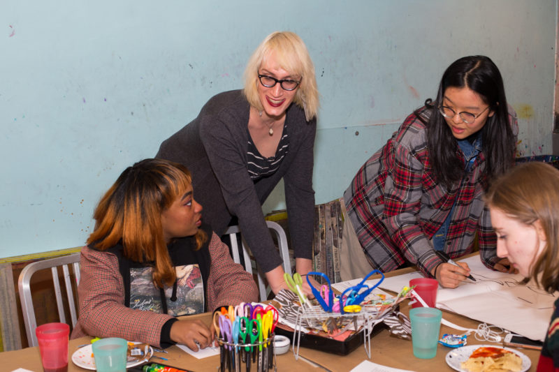 Four women are at a table. Two are standing next to the table and two are sitting. One has a black marker in her hand. There are art making supplies on the table.