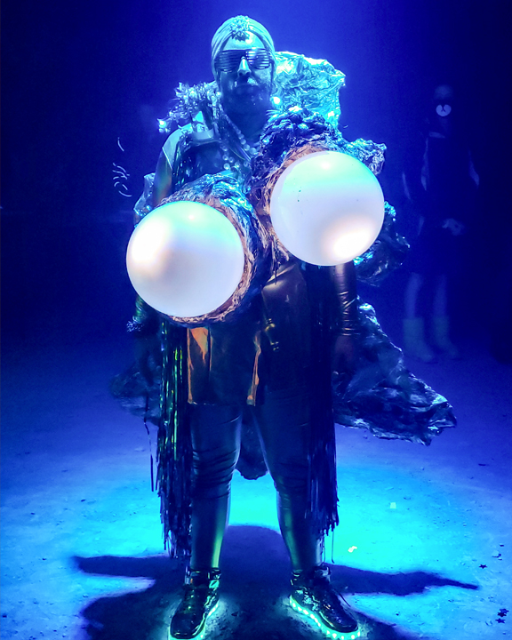 A person bathed in blue light wears shutter shades and light up shoes while holding two large white orbs.