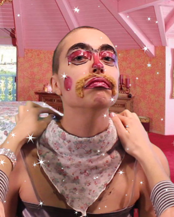 Person with a shaved head, theatrical makeup, and a mustache, wears a pink bandana while sitting in a pink room.
