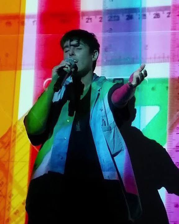 Person bathed in multicolored light speaks into a microphone with their eyes closed and left arm extended.