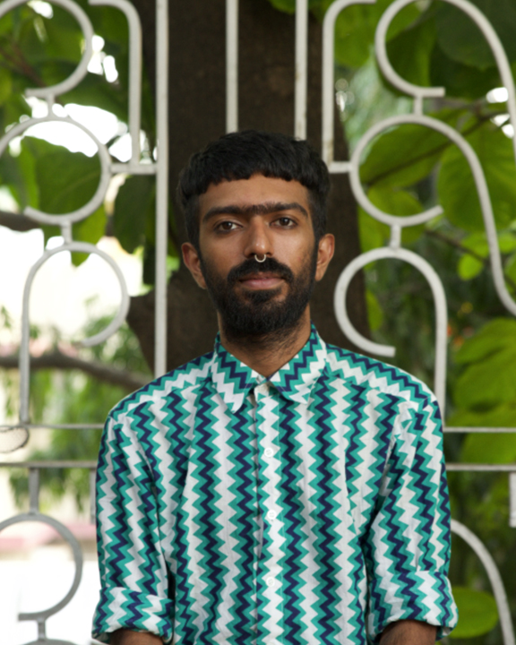 Headshot of a person in a patterned shirt with a beard and septum piecing in front of a white gate and foliage.