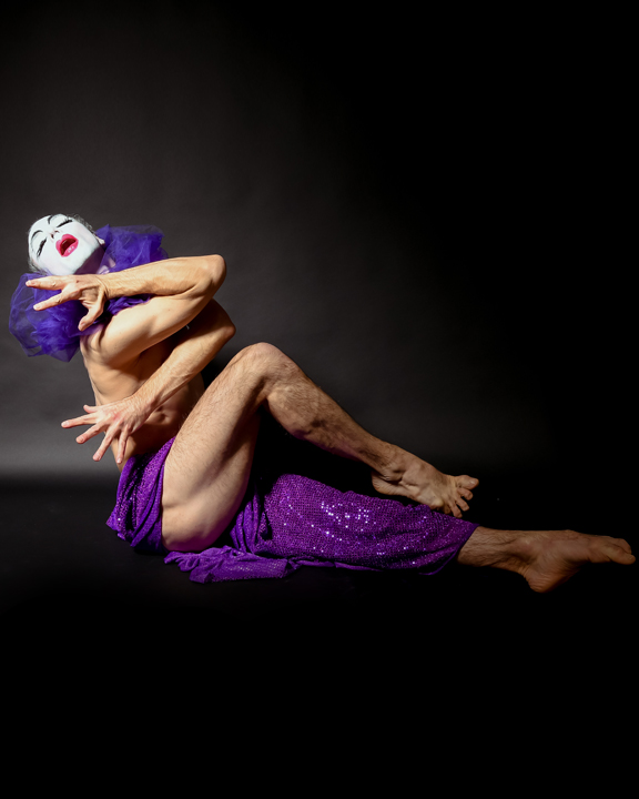 Person with blue hair and white face makeup contorts their body in a seated dance pose against a black background.