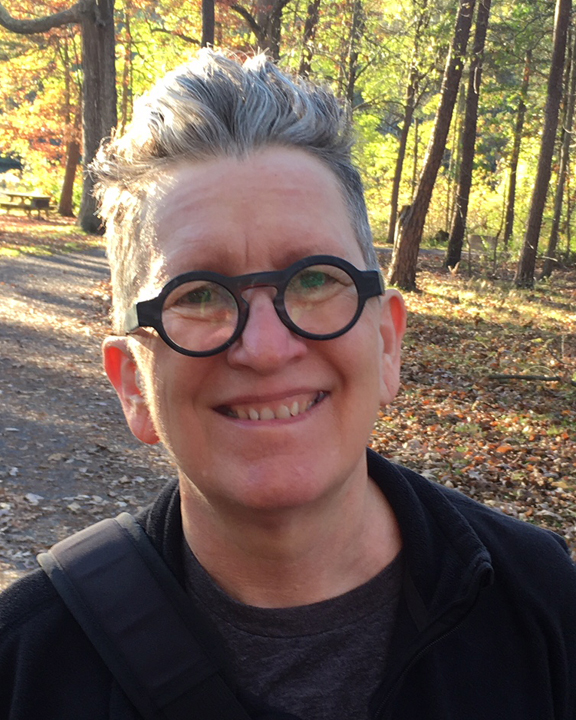 Headshot of a smiling person with short hair and round glasses in front of a sunny forest.