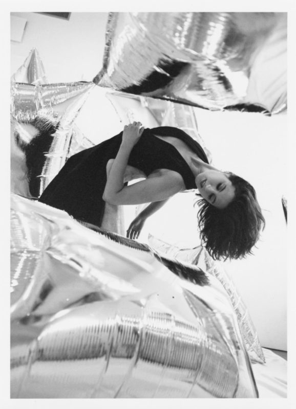 A black and white photo of a person in a black dress with long, dark hair, who is bending backwards in a room full of silver balloons.