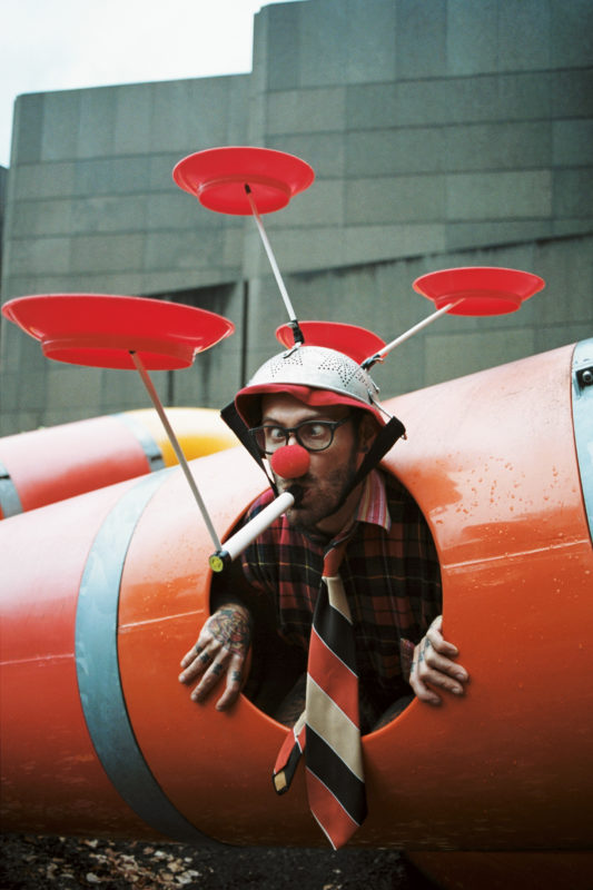 A person's head and hands are sticking out of a small hole. Their eyes are crossed and they have a long whistle in their mouth while balancing red plates on sticks that are stuck in a strainer on his head.