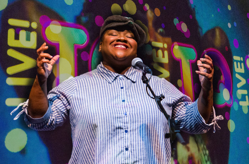 A performer smiles with their eyes closed and hands raised in front of a colorful background that reads “TQ Live!”