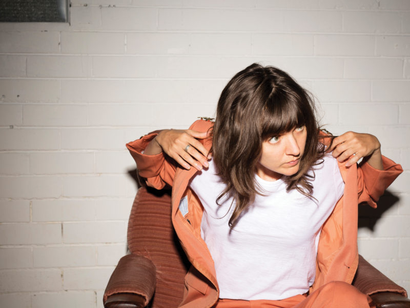 A person sits on a chair in front of a white brick wall. They are wearing a white shirt, orange pants, and an orange blazer. They are looking towards the right of the frame.