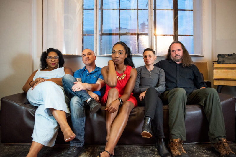 A group of five diverse individuals sit facing forward on a black couch with windows in the background.