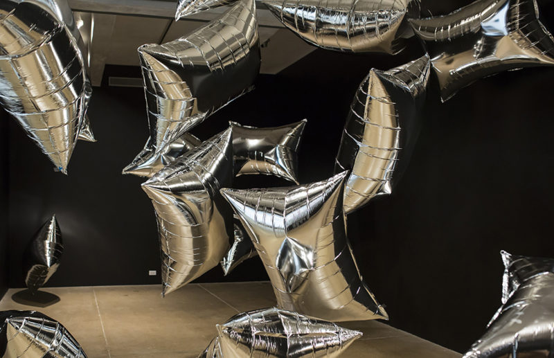 Helium and oxygen filled reflective silver Mylar pillows in a gallery space with black walls.