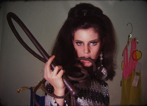 A film still of a person with long, brown hair looking at the camera while holding a whip in their right hand. Part of the whip is in their mouth.