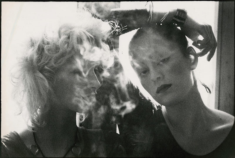 Black and white photograph of two people standing next to each other. There is smoke in front of their faces. The person on the left has light hair and is looking to their left. The person on the right has dark hair and has their right wrist, which has many bracelets on it, on top of their head while looking towards the camera.