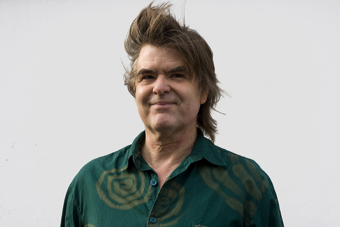 A person with a green button up shirt smiling at the camera while his hair is blowing in the wind