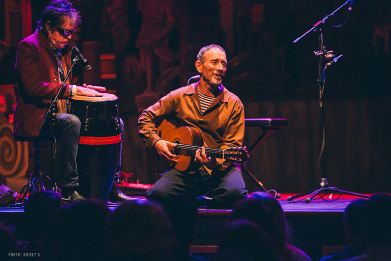 A person sits on a stage in front of a crown playing guitar. Another person is behind him sitting on a stool playing a bongo drum.