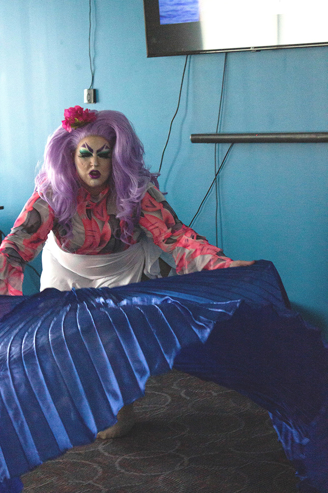 A performer stands in front of a blue wall. They have a full face of heavy, drag makeup. Their long, lilac-colored hair has a large red flower sitting in it. They are wearing a pink, green, and silver patterned long sleeve top, and leaning forward to twirl a flowing blue cape through the performance area.