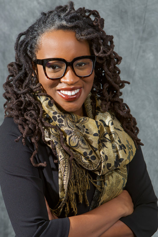 A photograph of a person with long, curly hair, wearing glasses, a decorative scarf, and a gray shirt smiles towards the camera.