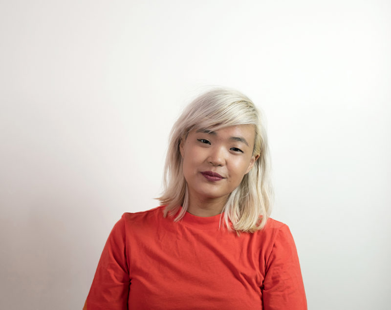 A person wearing red with light colored hair stands in front of a white background smiling towards the camera.