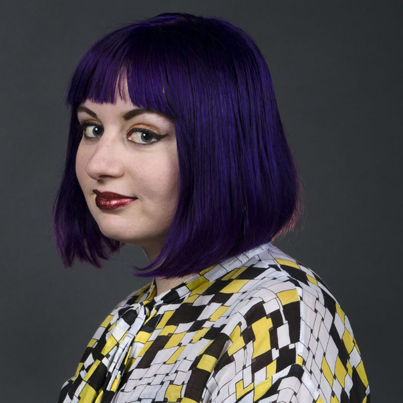 A person with short, purple hair smiles towards the camera.