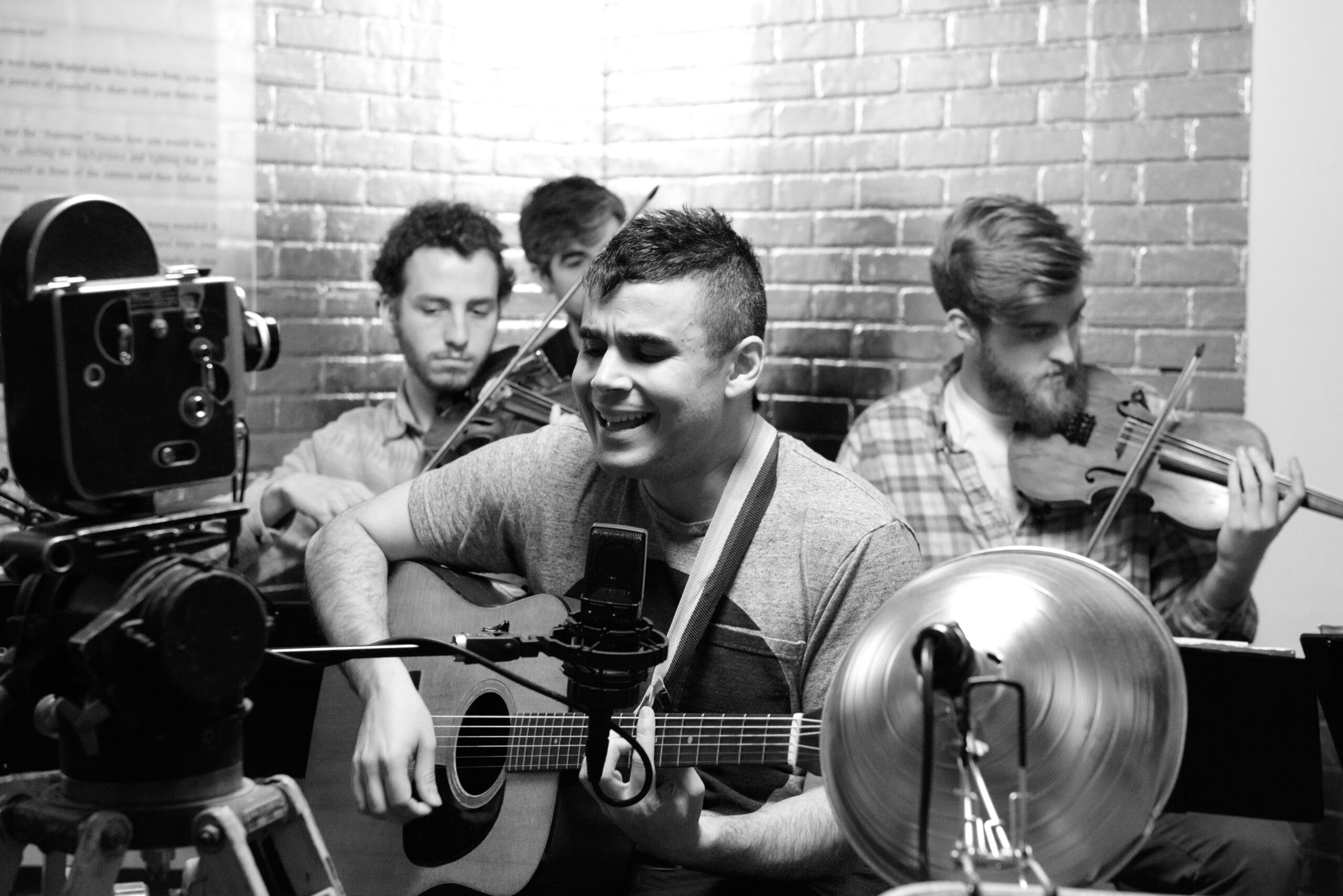 Man seated in foreground while playing guitar, three other musicians sitting behind him.