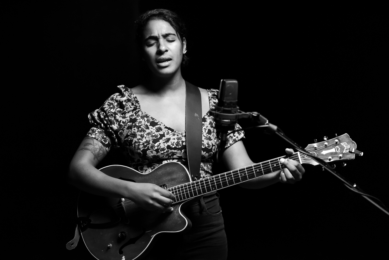 Woman playing an acoustic guitar stands at a microphone, her expression communicating that she is mid-song.