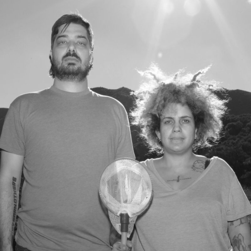 A tall man stands side-by-side with a shorter woman holding a butterfly net in a pose mimicking Grant Wood's American Gothic portrait.