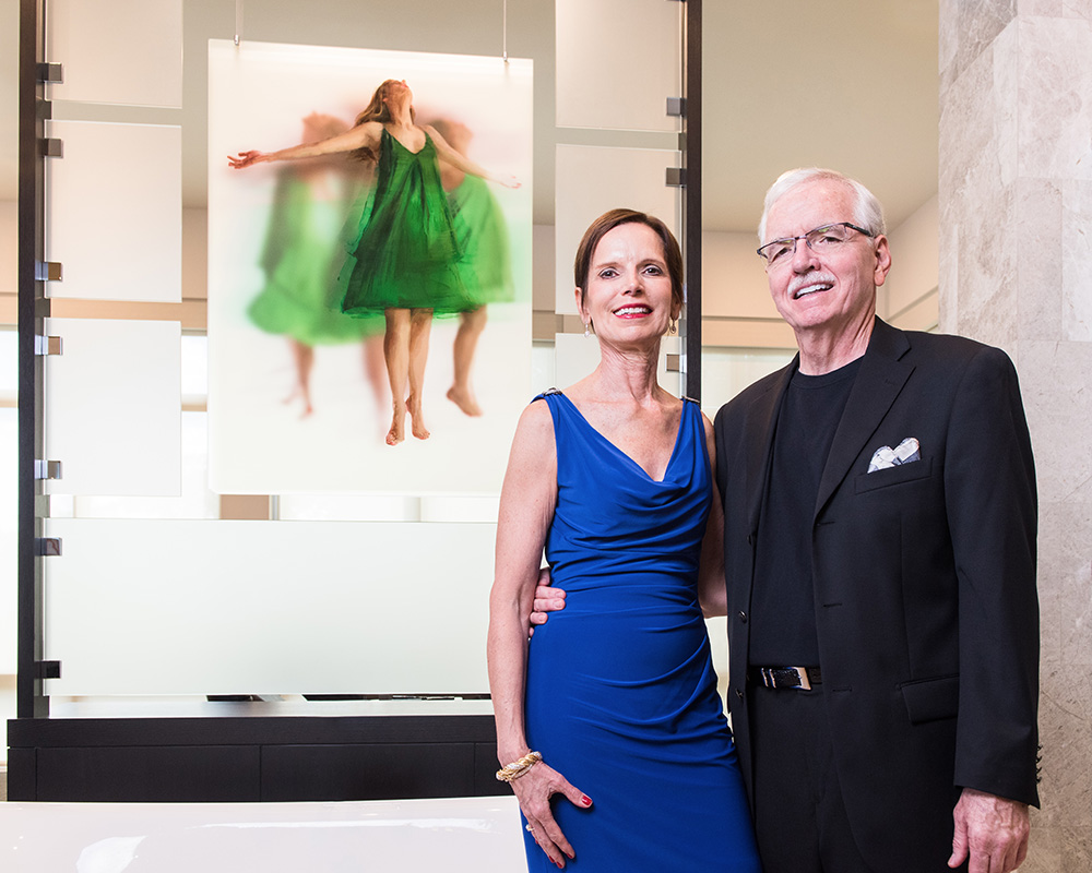 A person wearing a blue dress with short brown hair and a person wearing glasses and a black suit stand in front of an artwork depicting a person in a green dress with their arms raised up.