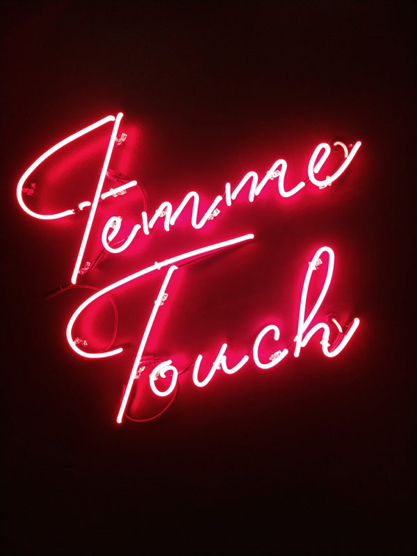 A reddish orange neon sign on a dark purple wall, with the words Femme Touch written in cursive.