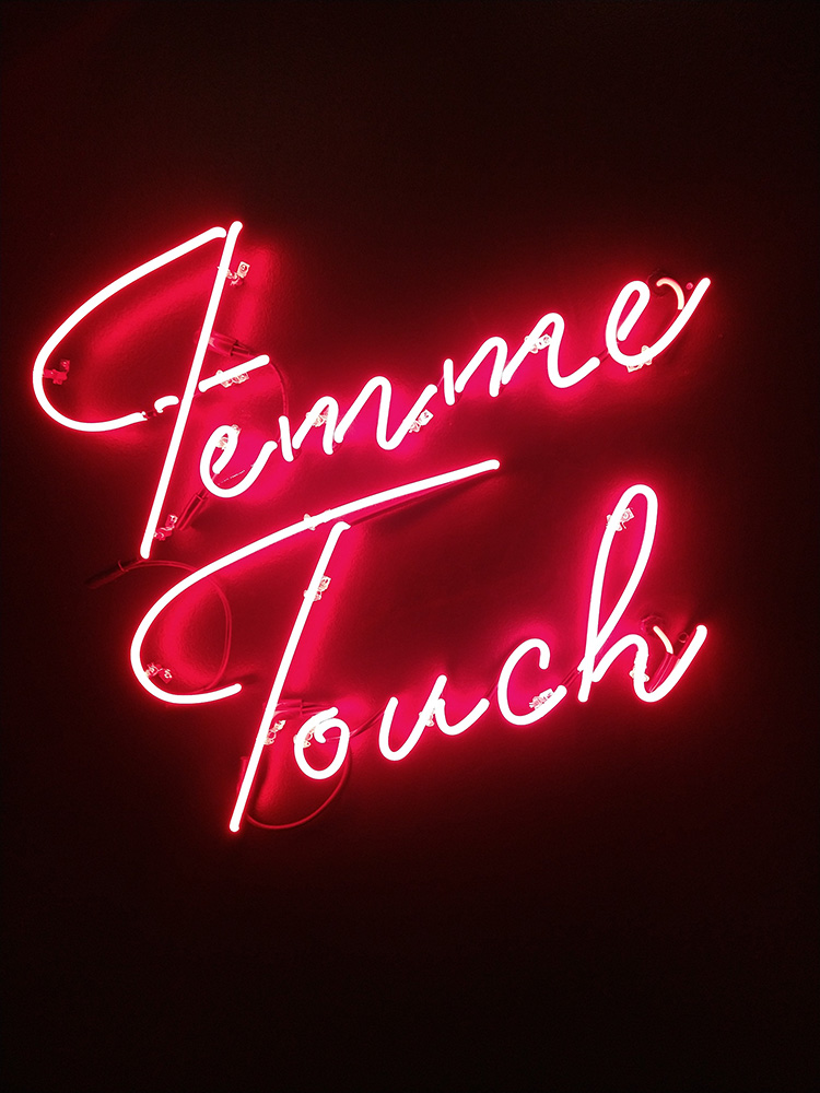 A reddish orange neon sign on a dark purple wall, with the words "Femme Touch" written in cursive.