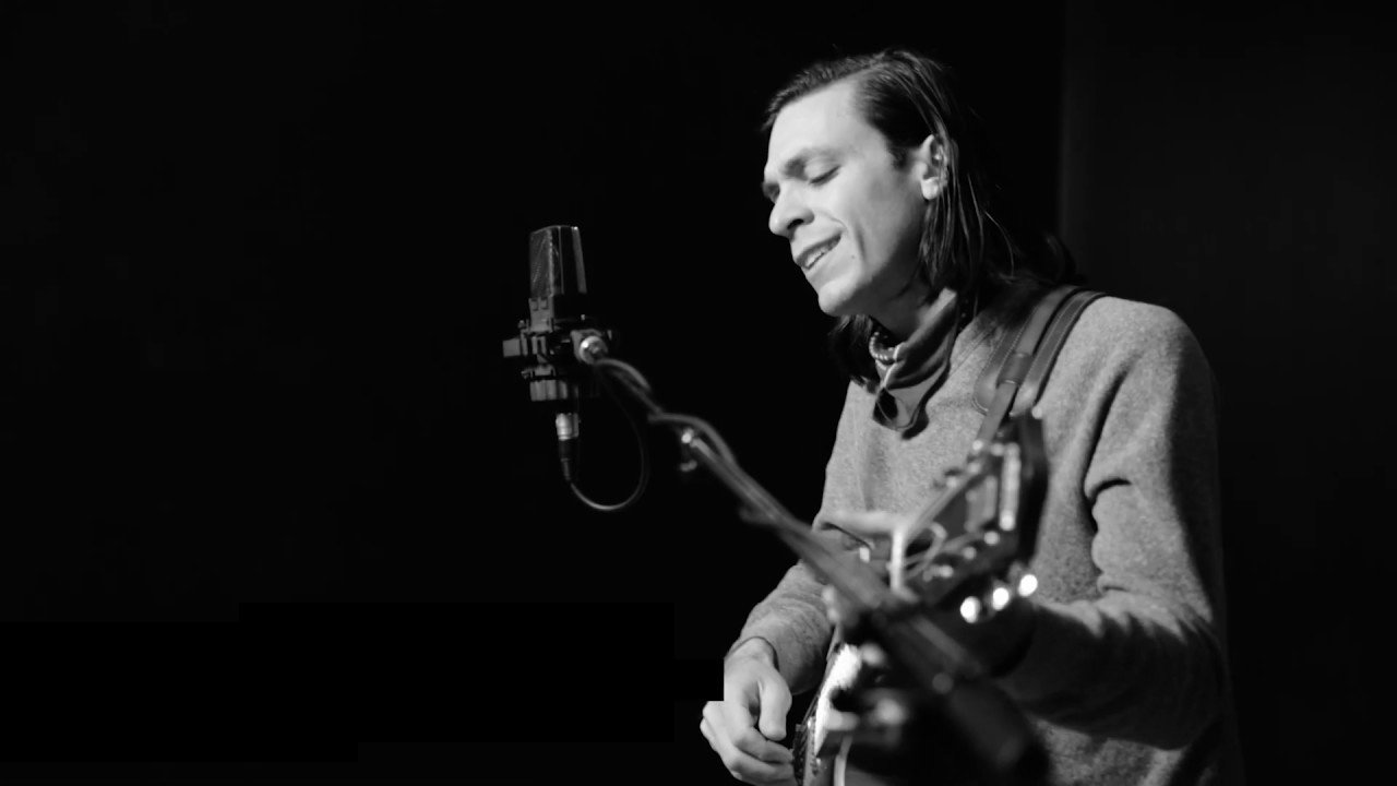 Man with long dark hair stands at a microphone while playing guitar.