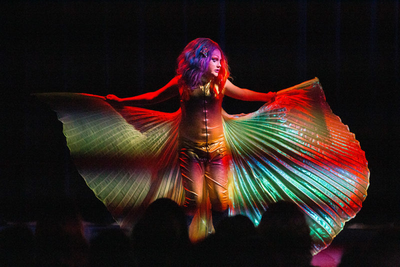 A drag performer is dramatically lit on a dark stage. They have a sparkly gold jumpsuit, multicolored hair, and a in their outstretched arms they are holding a flowing, sheer cape behind them. The lighting creates a rainbow effect when it catches the cape, and they are gazing off to the right, absorbed in their performance. Heads of audience members are silhouetted at the bottom of the frame.