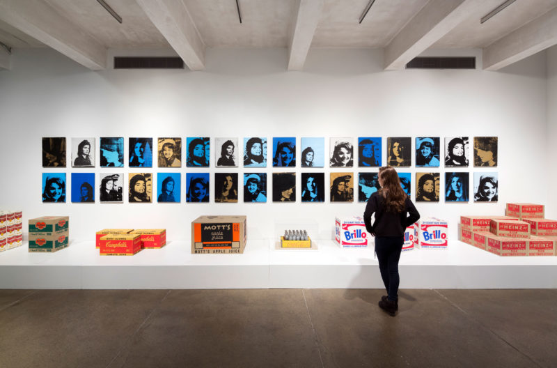 A woman with brown hair dressed entirely in black faces a display of Andy Warhol’s works. Two rows of small, screen-printed portraits are hung on a white wall. Some of Warhol’s sculptural work-- including reproductions of Campbell’s boxes, Brillo boxes, and a tray of silver Coca-Cola bottles-- sit on a low platform in front of the portraits.