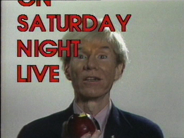 A video still of a person holding an apple and looking towards the camera. A Saturday Night Life graphic in red, capital letters appears in the foreground.