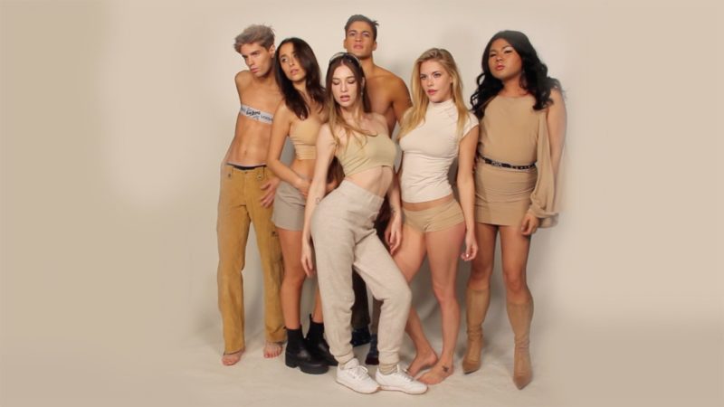 Six people stand in a group, each posing as if for a fashion shoot. Visible from head-to-toe, they are all wearing outfits of varying shades of beige, and are standing against a solid beige background.