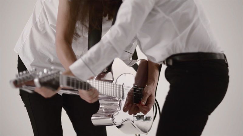 Two people in white shirts and black pants face each other, each playing one side of a guitar that has two sides and two necks.