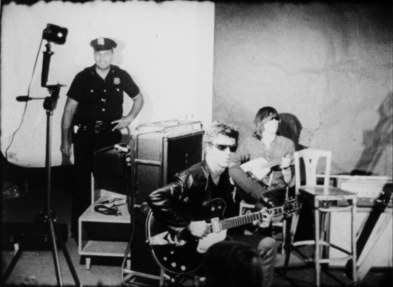 Black and white film still of two members of the Velvet Underground sitting in a studio with instruments in their hand. There is a police officer standing in the background.