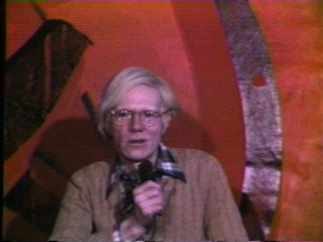 Film still of a person holding a mic in front of a red background. He is shown here with a pair of round glasses, wearing a black and white plaid shirt underneath a brown sweater.