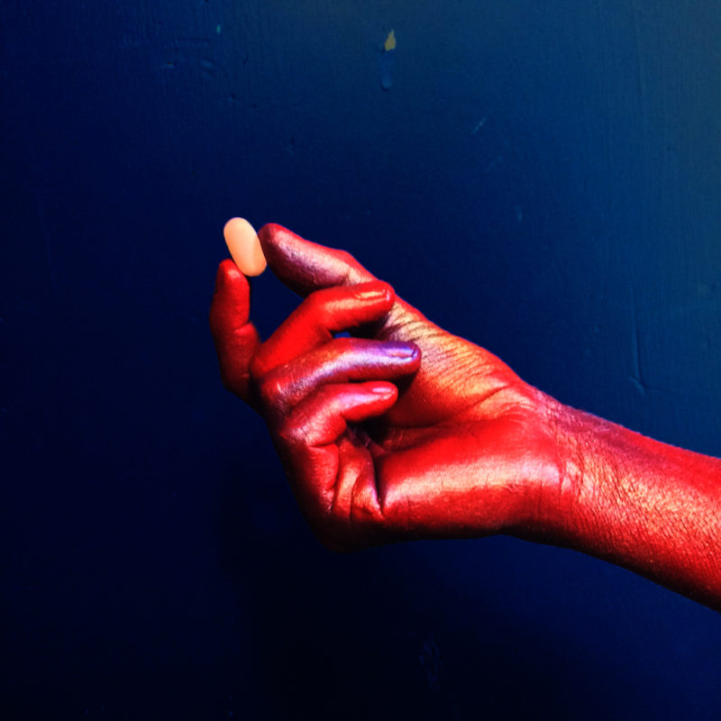 A hand painted in red holding a yellow pill in front of a dark blue background.