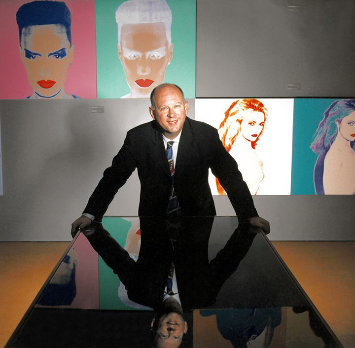A person in a suit is leaning over a black table in a gallery with artworks hanging behind him on a gray wall.