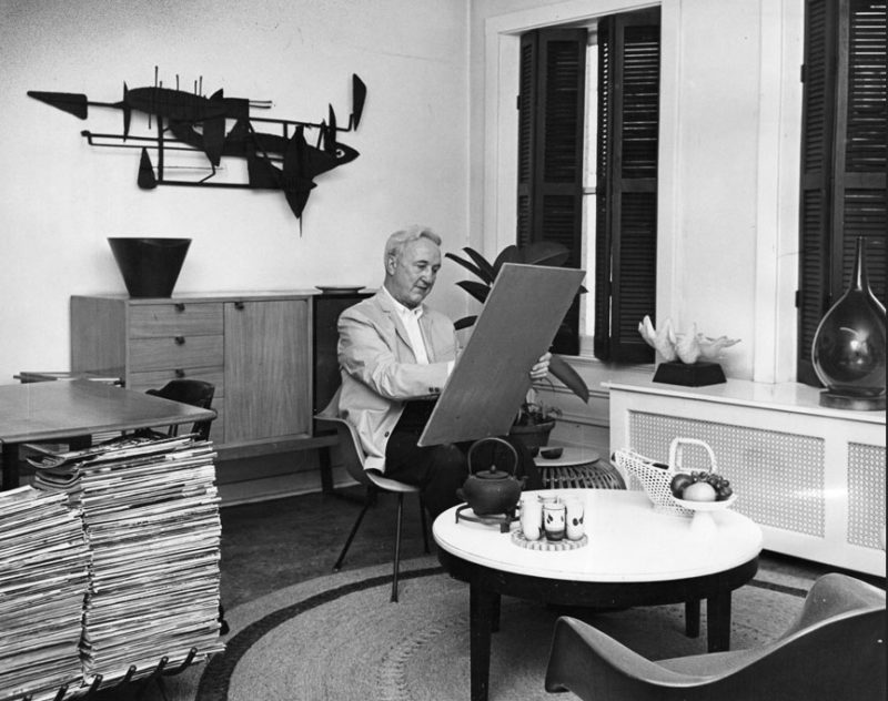 Man sits in the center of his living room, art board in hand, drawing.