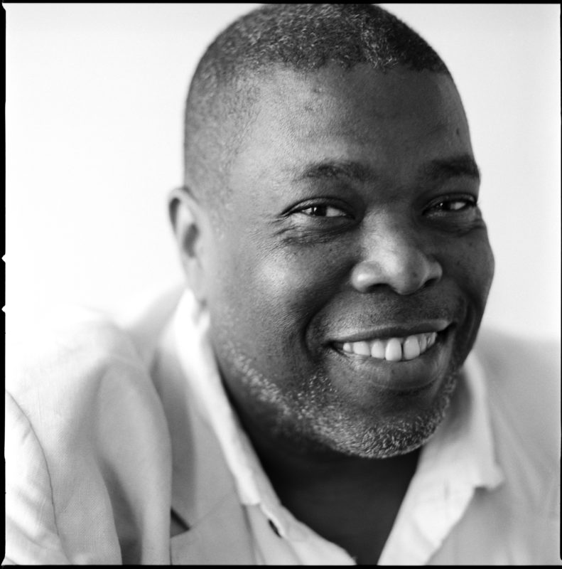 Writer and theater critic Hilton Als, wearing a light colored blazer and dress shirt, smiles at the camera.