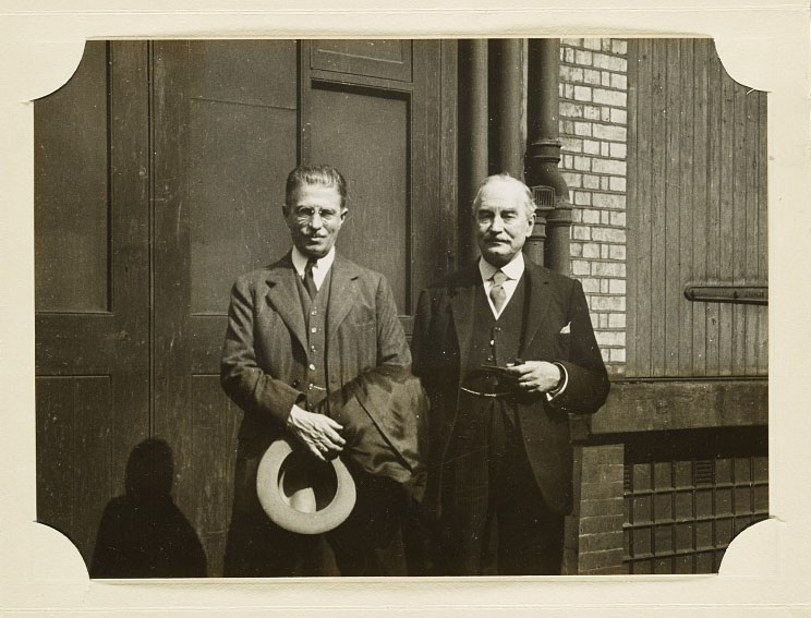 Two men, one holding a hat in his hands, stand side by side.