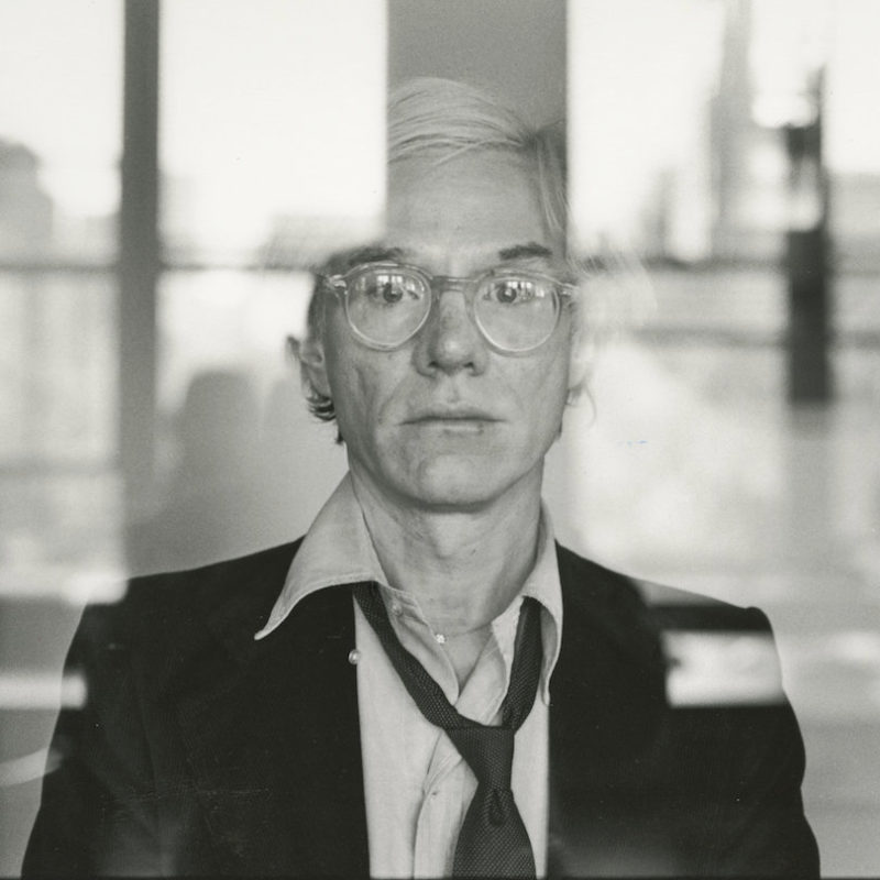 Warhol, standing behind a glass window, looks out at the viewer.