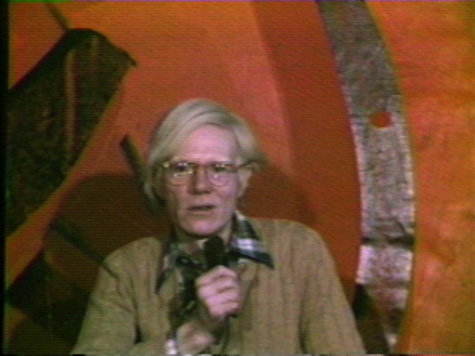Andy Warhol holding a microphone and speaking while looking directly into camera